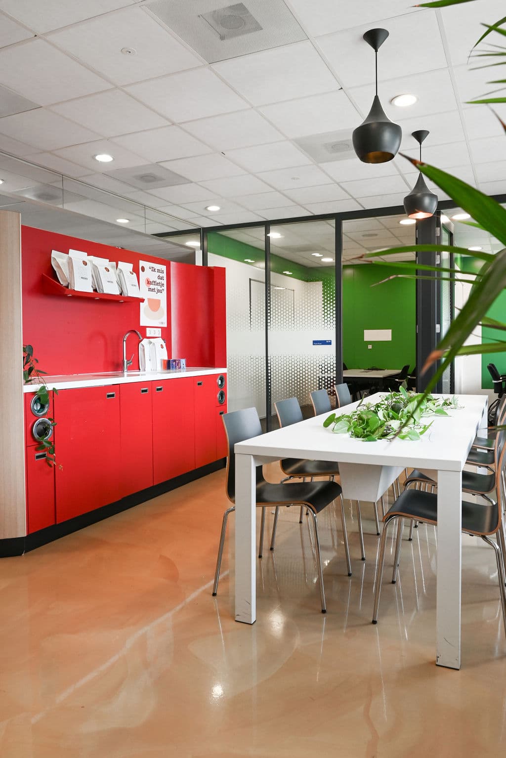 Modern office kitchen with red cabinets, a white table, gray chairs, and hanging black lights. Green plants add a touch of nature.