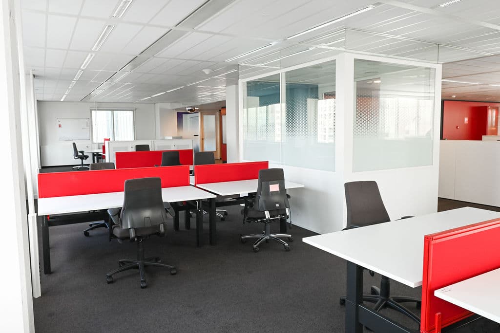 Modern office space with white desks, red dividers, and black chairs. Glass partitions and bright lighting enhance the open layout.