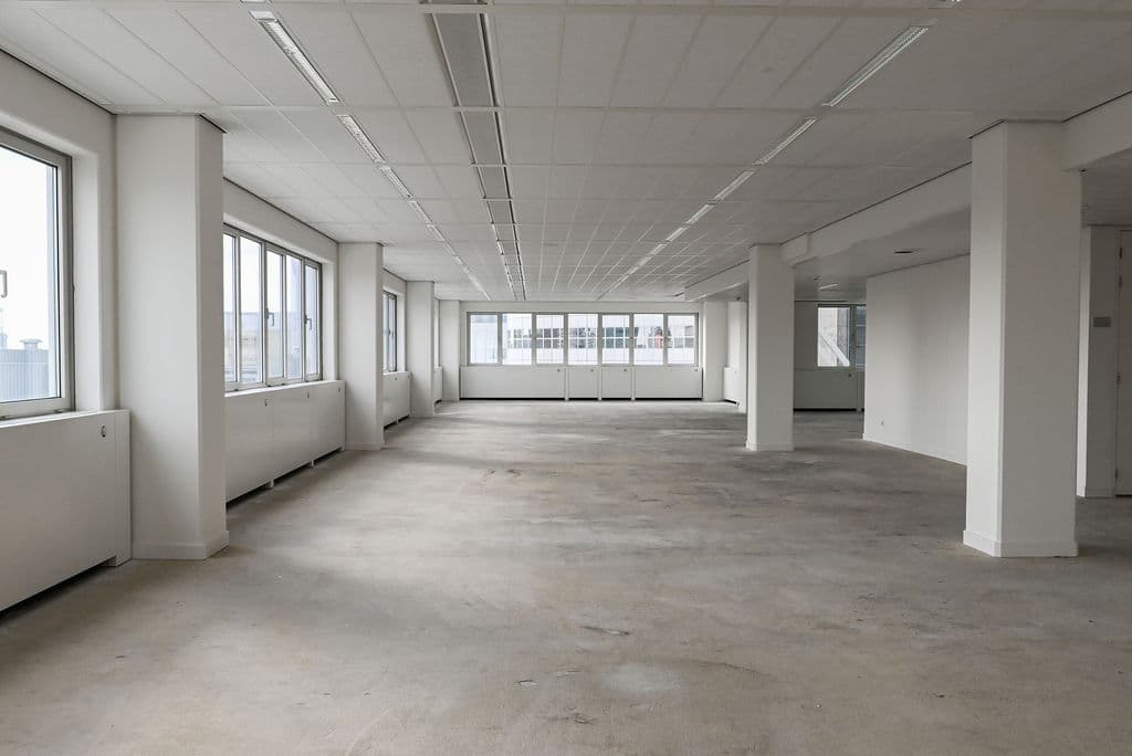 Empty office space with large windows, white walls, and exposed flooring. A person is walking in the distance near the entrance.