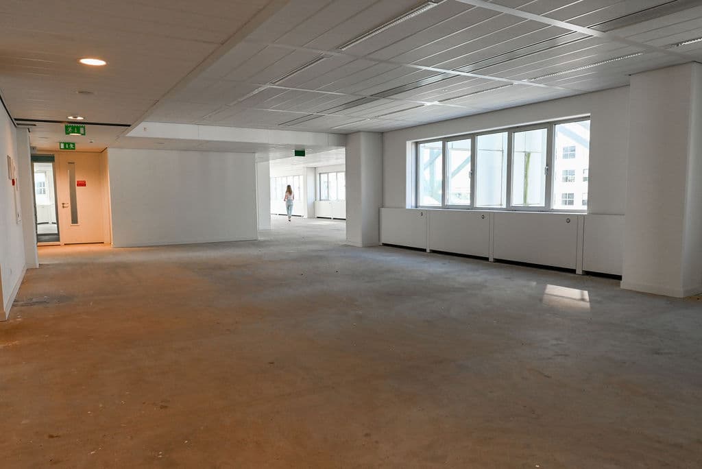 Empty office space with large windows, white walls, and exposed flooring. A person is walking in the distance near the entrance.