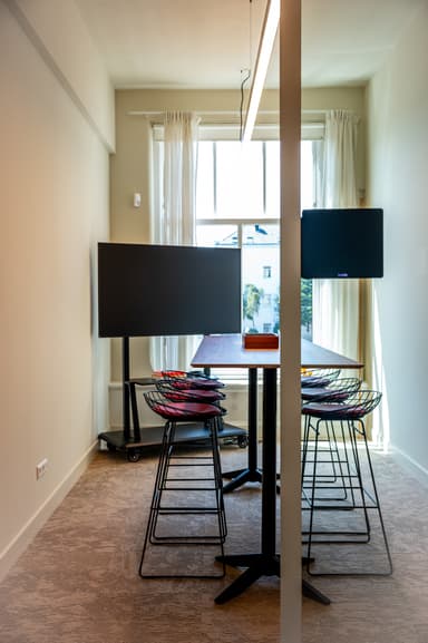 Modern conference room with a long table, wire chairs, two large screens, and a window with sheer curtains letting in natural light.