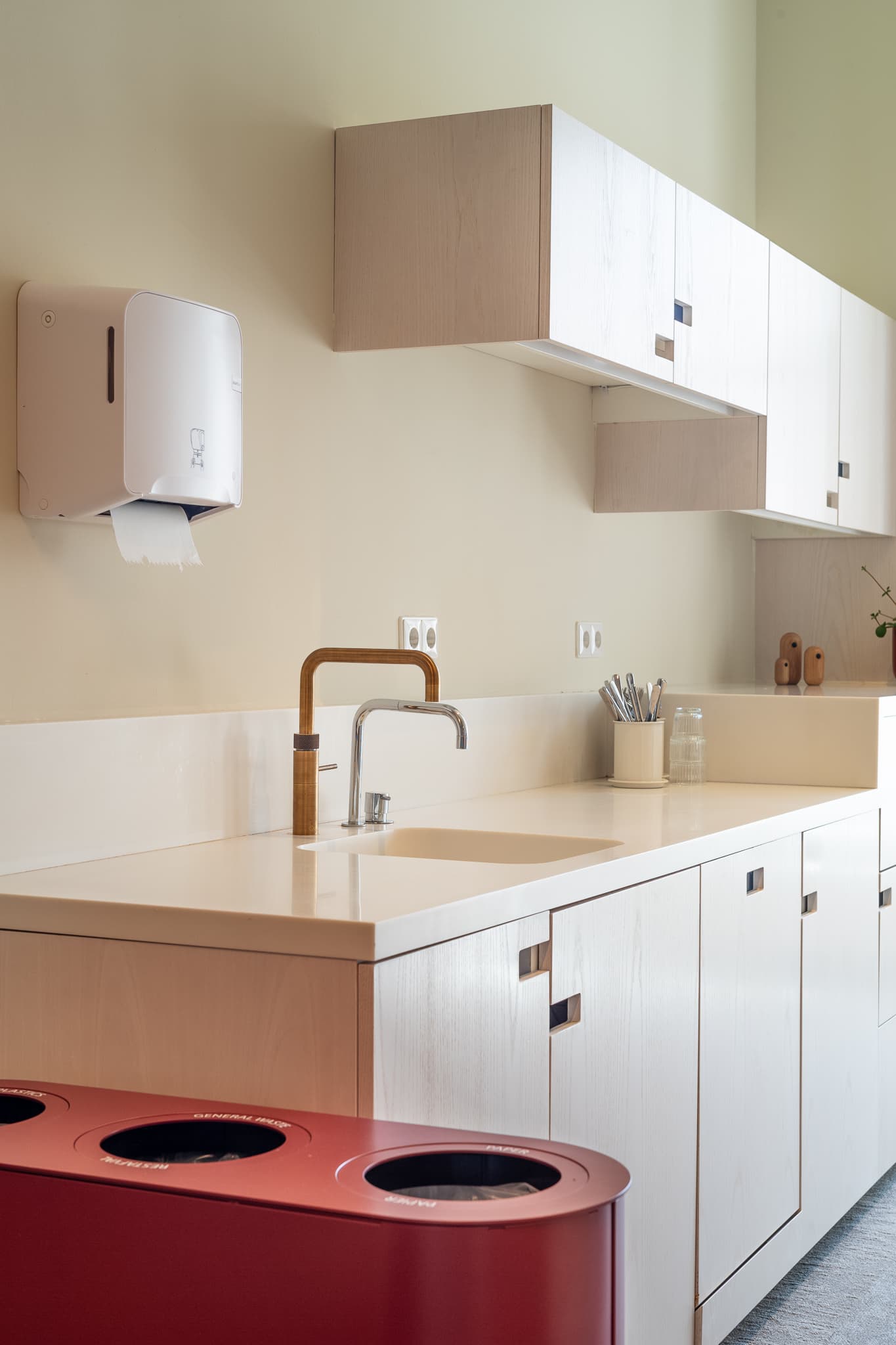 Modern kitchen with light wood cabinets, a white countertop, a stainless steel faucet, and a red recycling bin. Wall-mounted paper towel dispenser.