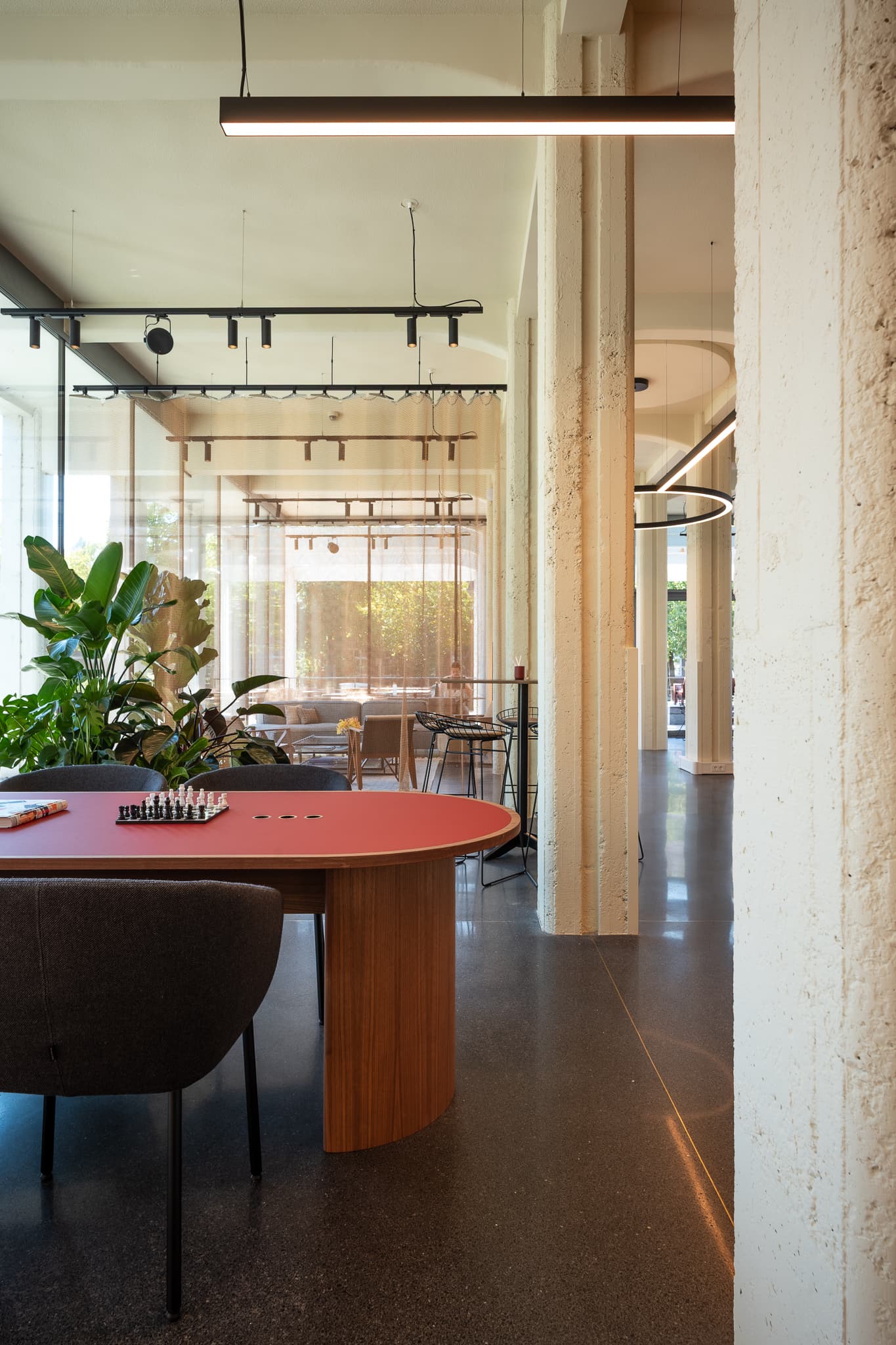 Modern interior with a red oval table set for chess, surrounded by chairs, large plants, and floor-to-ceiling windows with sheer curtains.