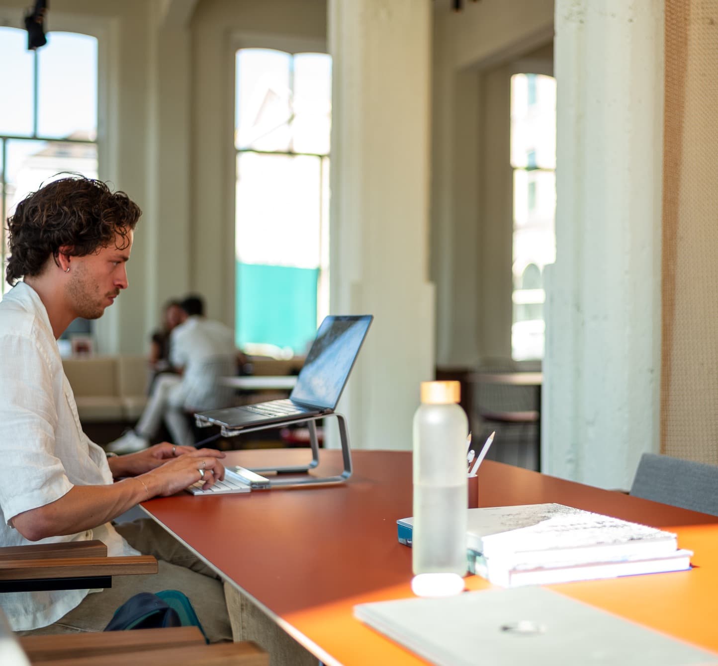 Man working on a laptop at a bright, modern office table with books and a water bottle. Background shows large windows and seating area.