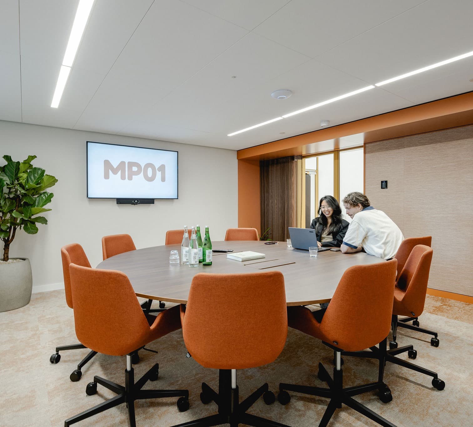 Two people work at a laptop in a modern conference room with orange chairs around a large round table and a screen displaying "MP01."