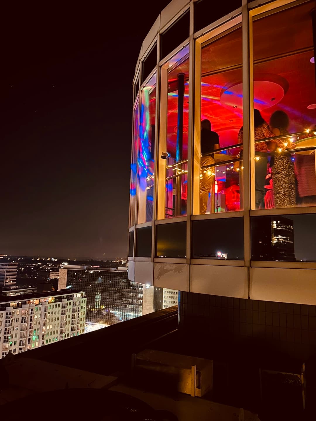 Nighttime rooftop bar scene with colorful lights and patrons inside a glass-walled space, overlooking a cityscape with illuminated buildings.