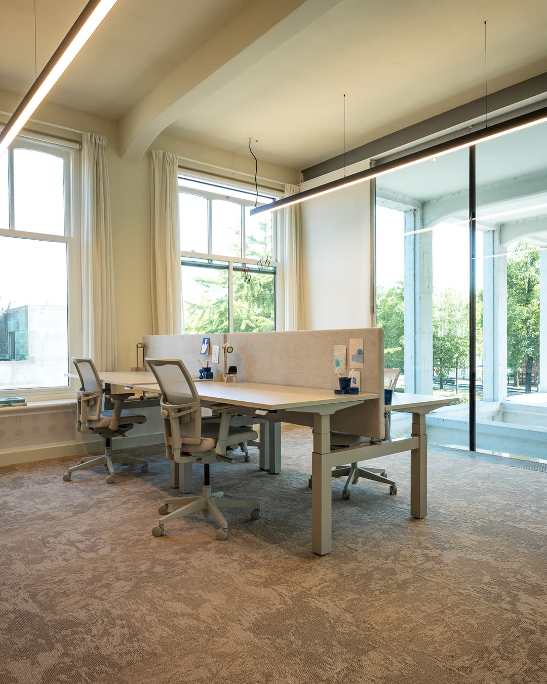 Modern office space with two workstations, ergonomic chairs, large windows, and glass walls overlooking greenery. Bright and spacious ambiance.