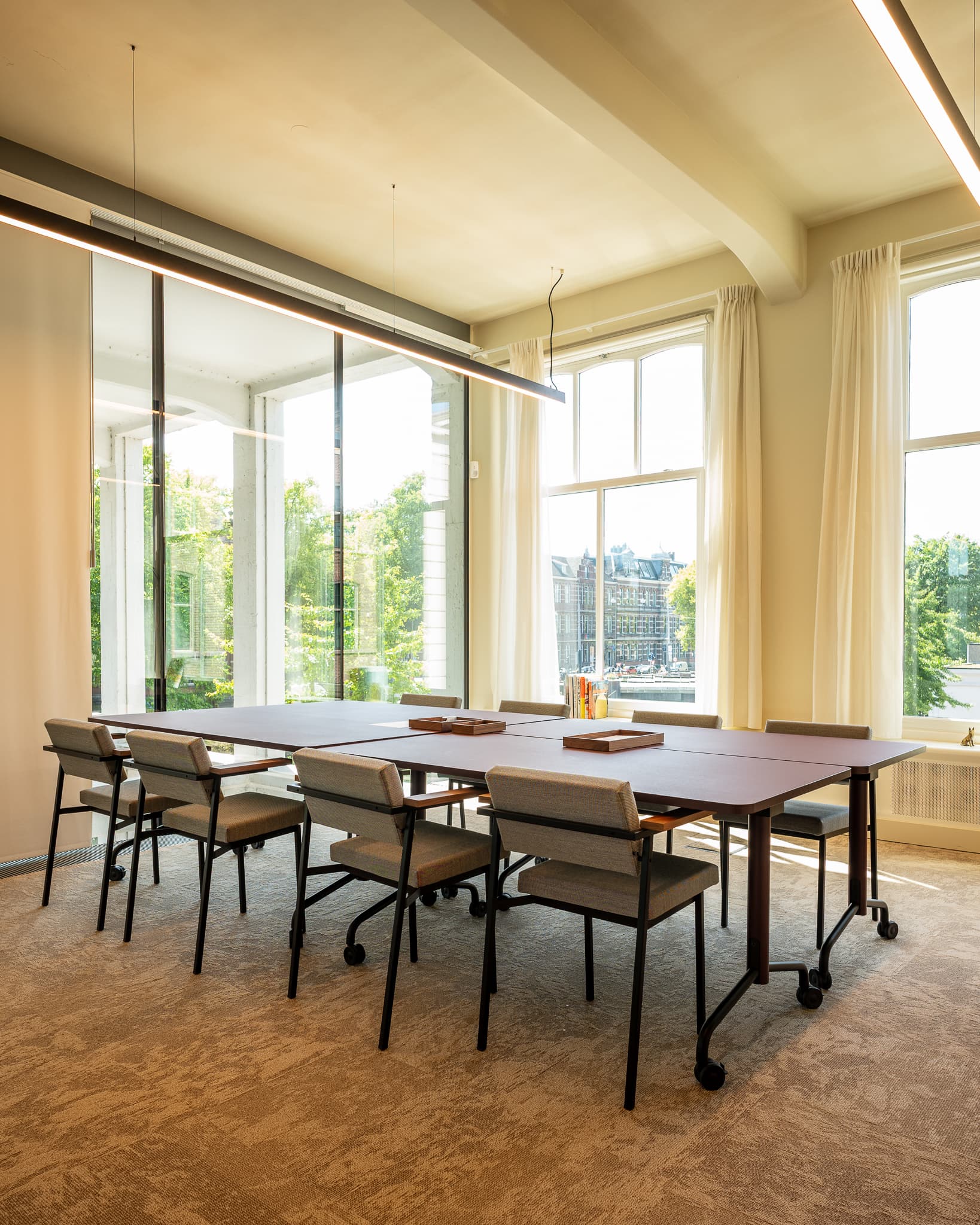 A modern conference room with a long table, chairs, large windows, and a view of greenery outside. Bright lighting and neutral tones.