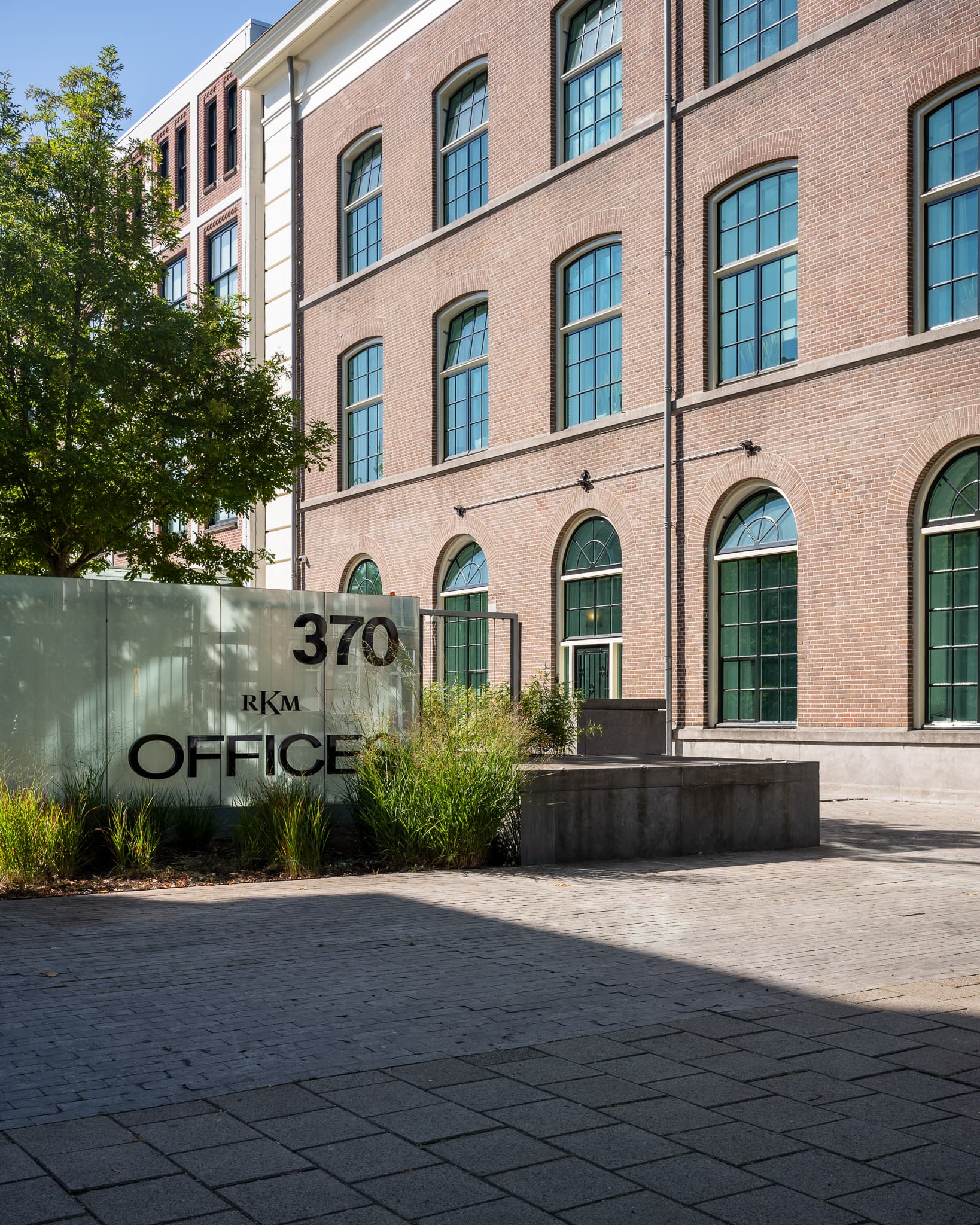 Brick office building with arched green windows, glass sign reading "370 KM OFFICE," and a tree in the foreground.