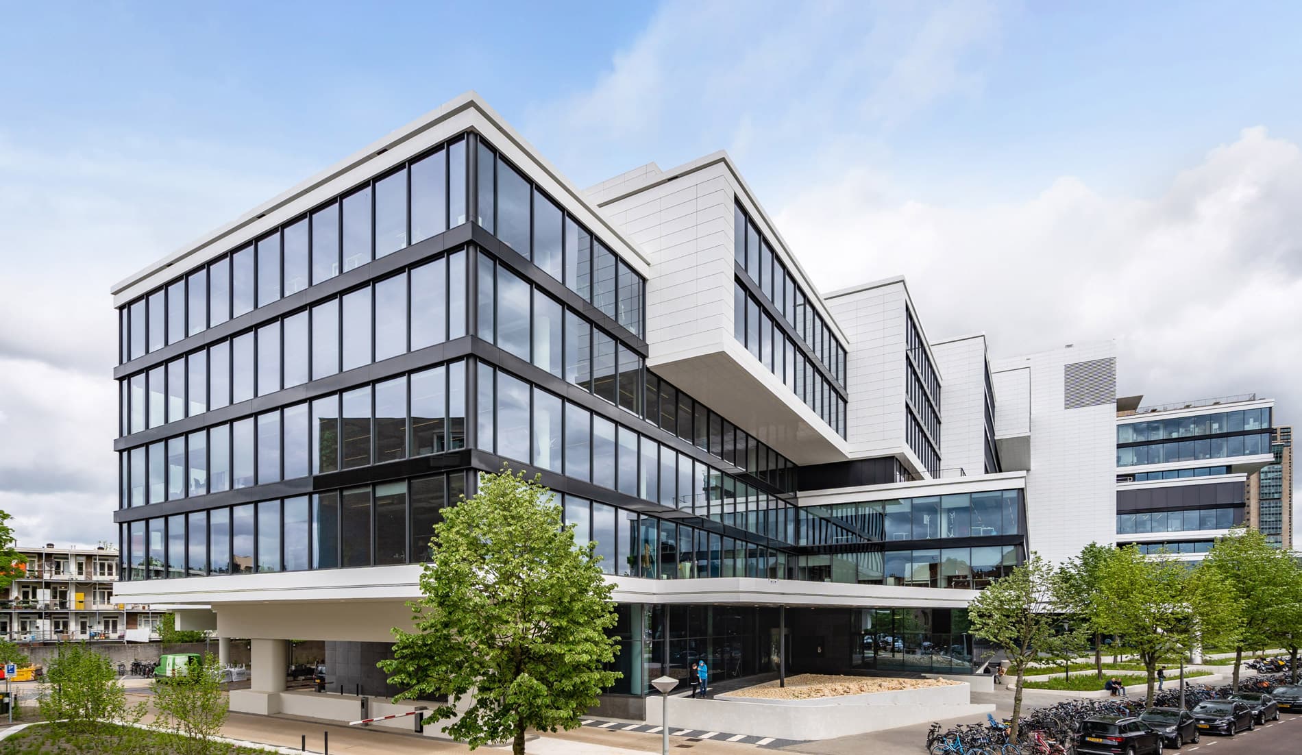 Modern office building with large glass windows, angular design, and surrounding greenery under a partly cloudy sky.