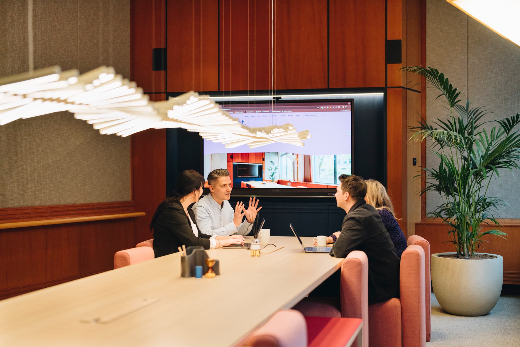 Four people in a modern conference room discussing at a long table with laptops and a large screen displaying a presentation.