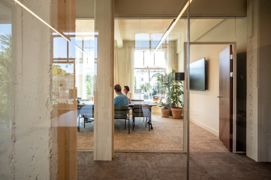 Modern conference room with a long table, chairs, large screens, and a window with sheer curtains letting in natural light.