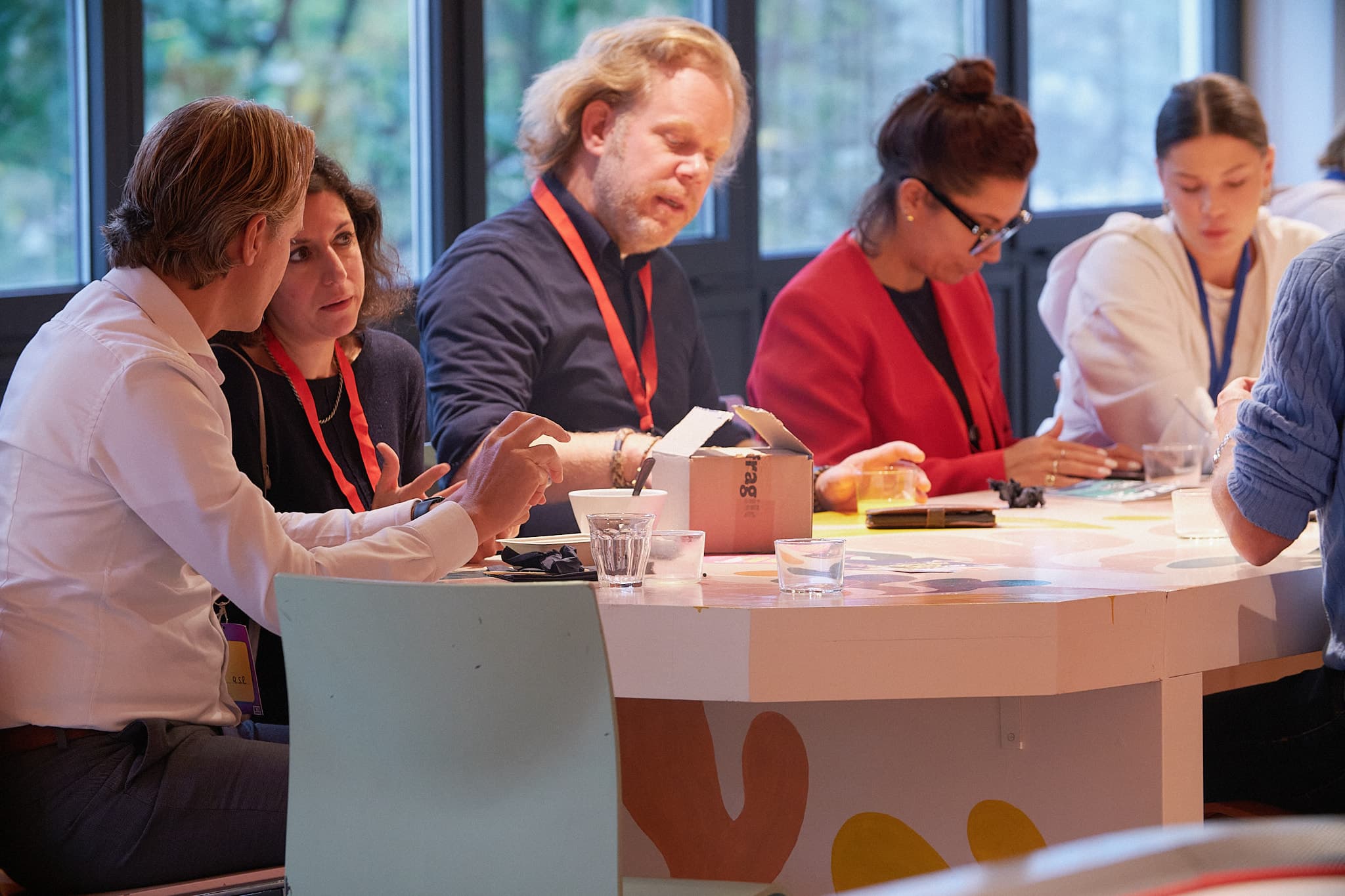 People sitting around a table engaged in discussion, with papers and drinks in front of them, in a well-lit room with large windows.