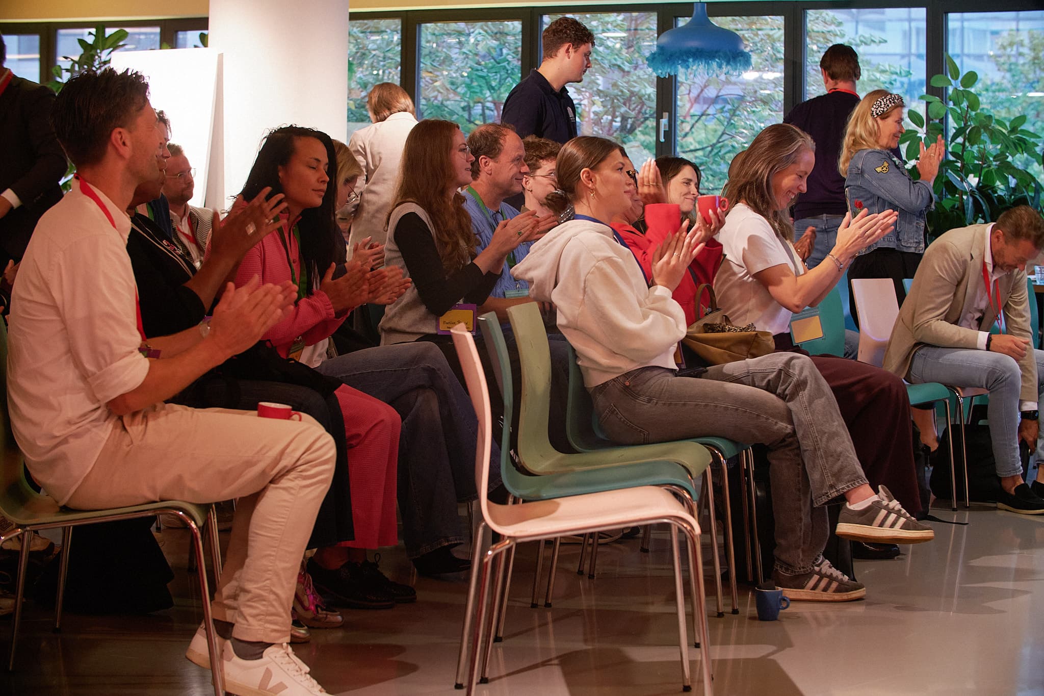 A group of people seated in a room, clapping and engaged, with some standing near large windows.