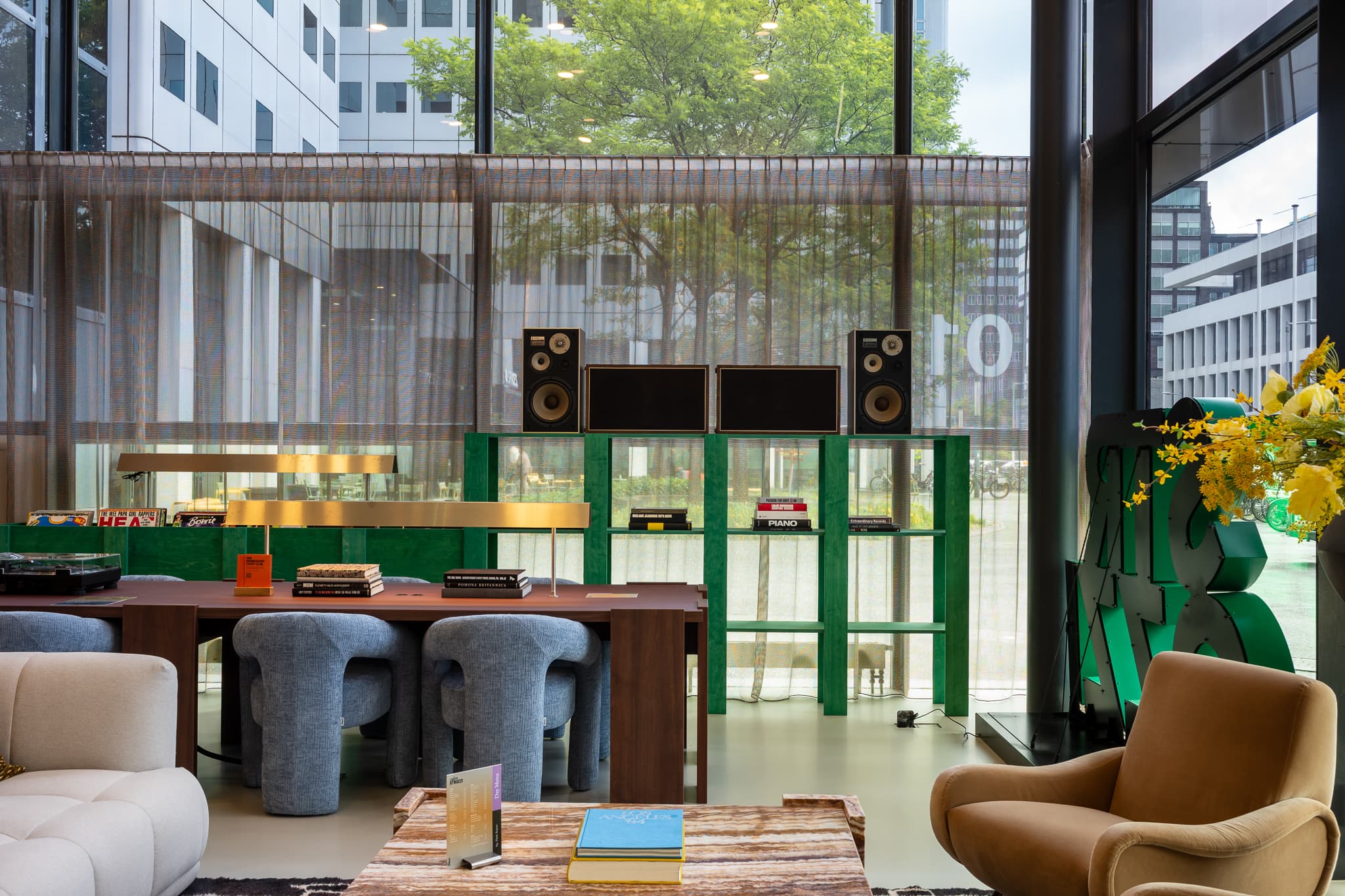 Modern lounge with plush seating, a wooden table, books, and speakers. Large windows reveal a view of urban greenery outside.