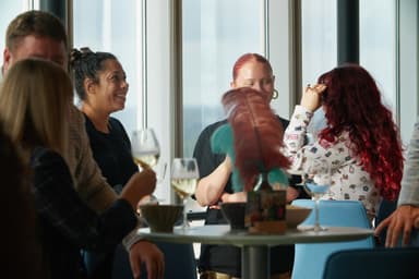 A group of people socializing around a table with drinks, near large windows.