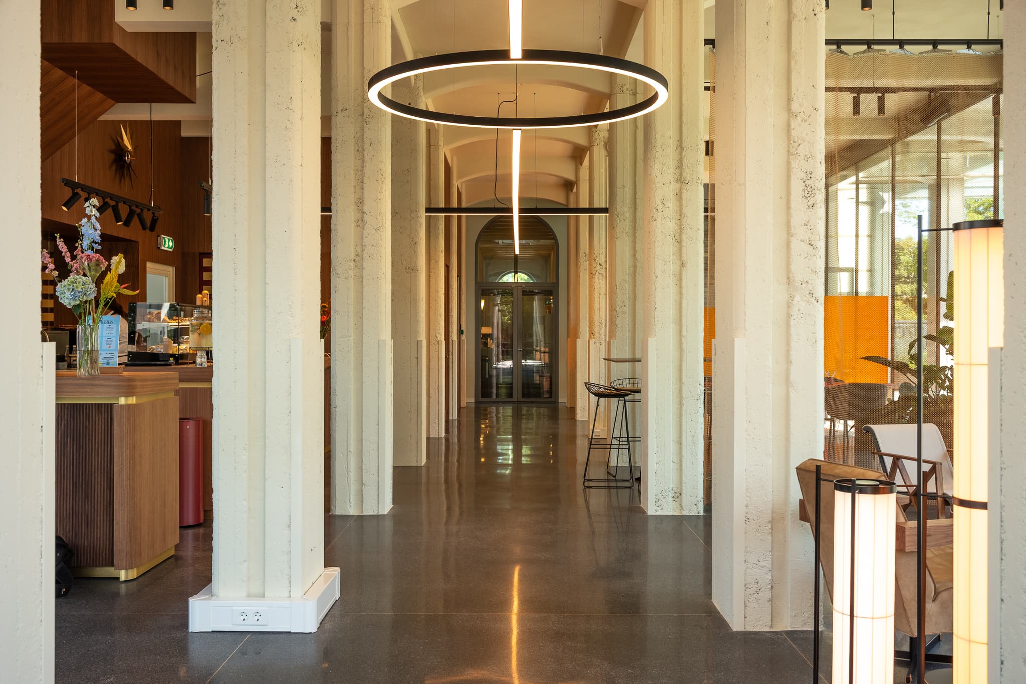 Modern interior with tall columns, circular ceiling lights, and a shiny floor. A café counter is on the left, and seating is on the right.