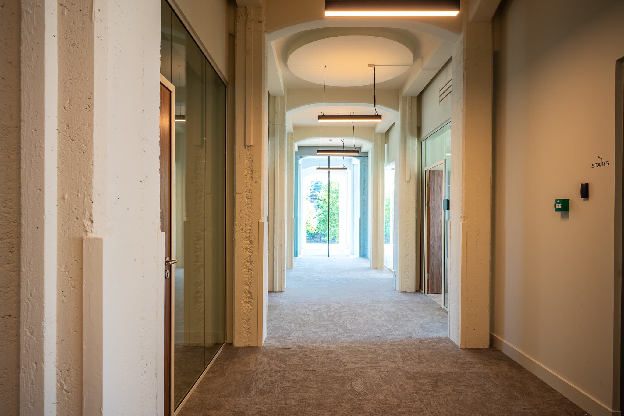 Modern hallway with textured white walls, glass doors, and ceiling lights, leading to a bright window with a view of greenery.