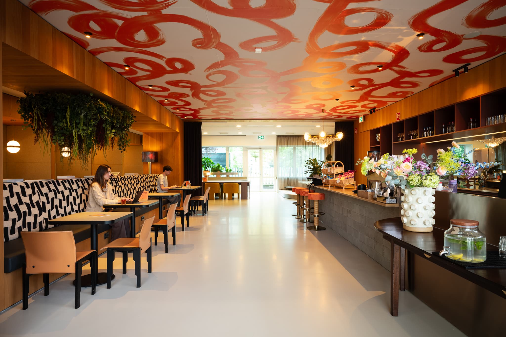 Modern cafe interior with bold red ceiling design, cozy seating, lush plants, and a decorative bar area with floral arrangements and refreshments.
