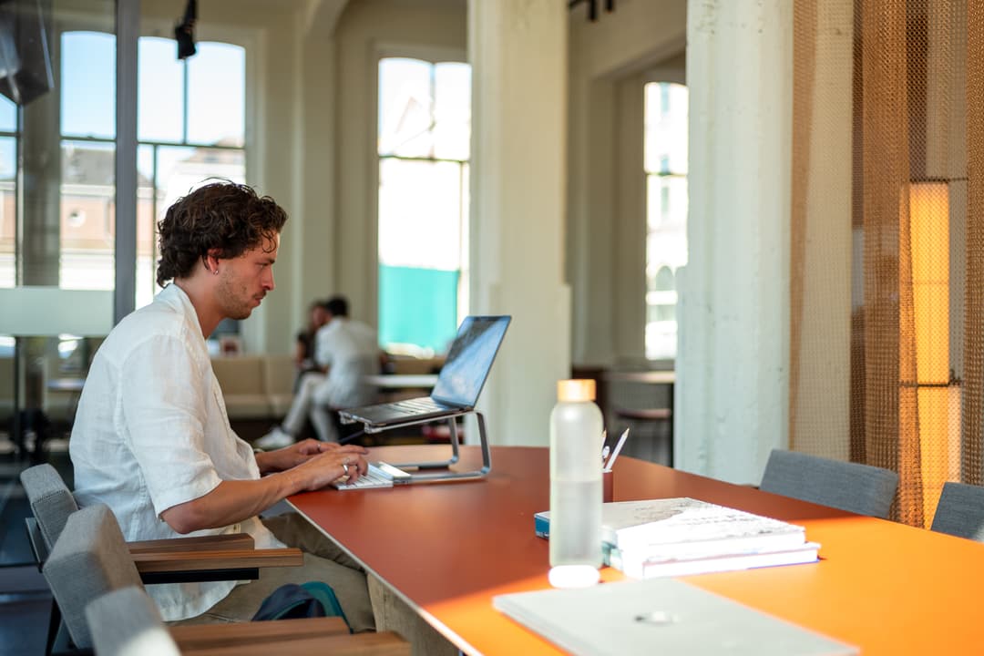 Man working on a laptop at a long table in a bright room with large windows. A water bottle and notebooks are on the table.