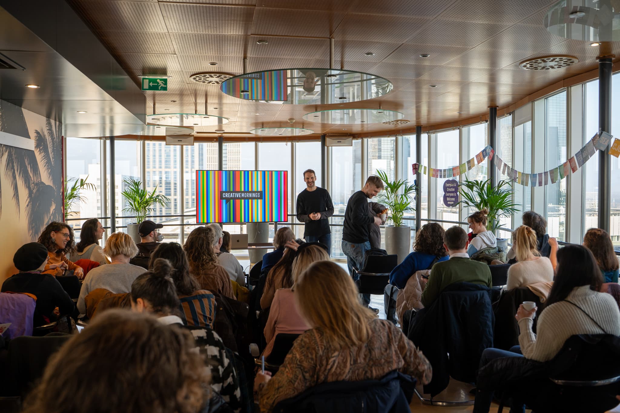 A speaker presents to an audience in a modern, bright room with city views and colorful presentation slides visible in the background.