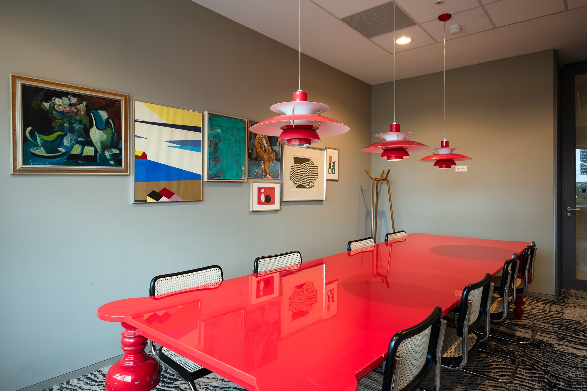 Modern meeting room with a long red table, black chairs, and red pendant lights. Artworks decorate the grey walls.