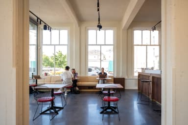 Modern cafe interior with large windows, minimalist decor, and a few people sitting at tables. Bright natural light fills the space.