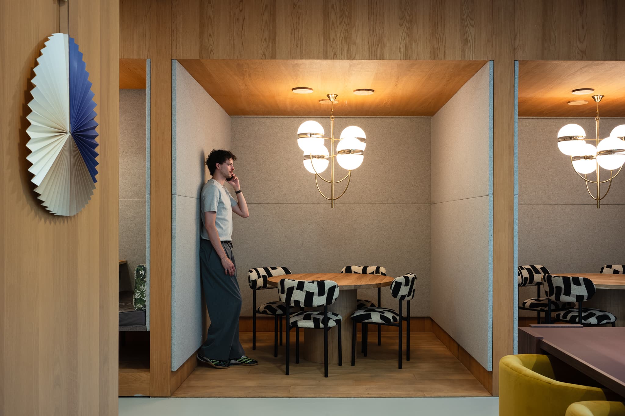 Man on phone in modern meeting booth with wooden table, patterned chairs, and chandelier; paper wall decoration on the side.