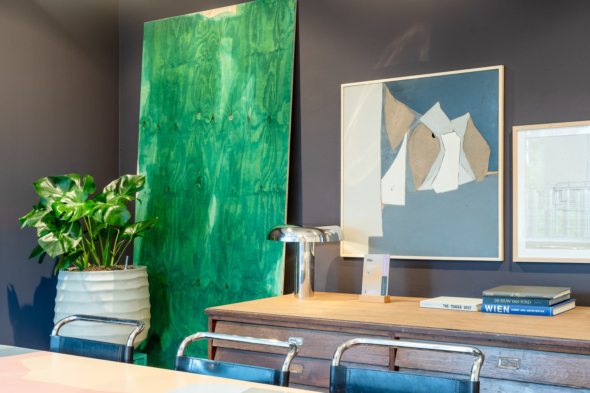 Office corner with a leafy plant, artistic wall panel, abstract painting, sleek lamp, and books on a wooden desk.