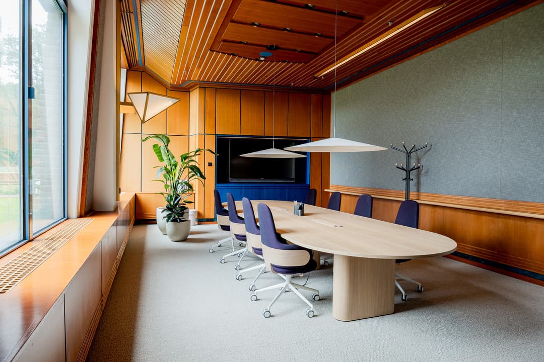 Modern conference room with a wooden oval table, purple chairs, large windows, potted plant, and ceiling lights. A screen is mounted on the wall.
