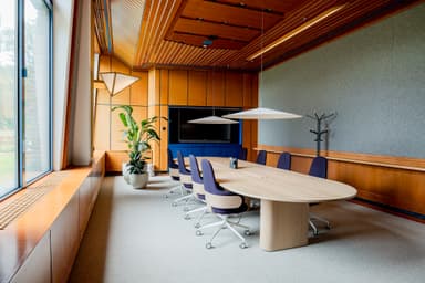Modern conference room with a wooden oval table, purple chairs, large windows, potted plant, and ceiling lights. A screen is mounted on the wall.