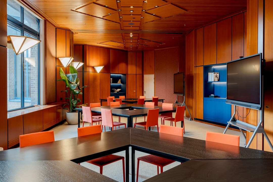 Modern conference room with wooden paneling, triangular tables, orange chairs, large windows, and digital screens. Bright, geometric lighting fixtures.
