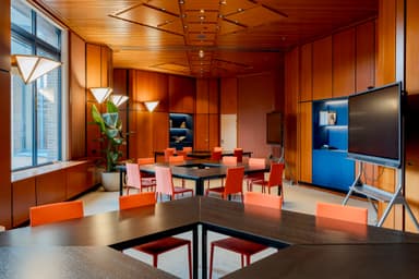 Modern conference room with wooden paneling, triangular tables, orange chairs, large windows, and digital screens. Bright, geometric lighting fixtures.
