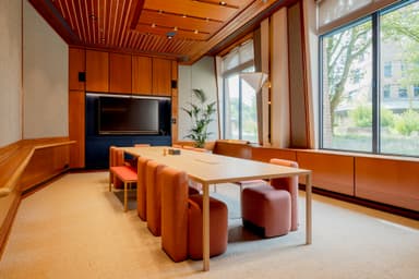 Modern conference room with a long wooden table, red chairs, a large TV, and large windows overlooking a green outdoor area.
