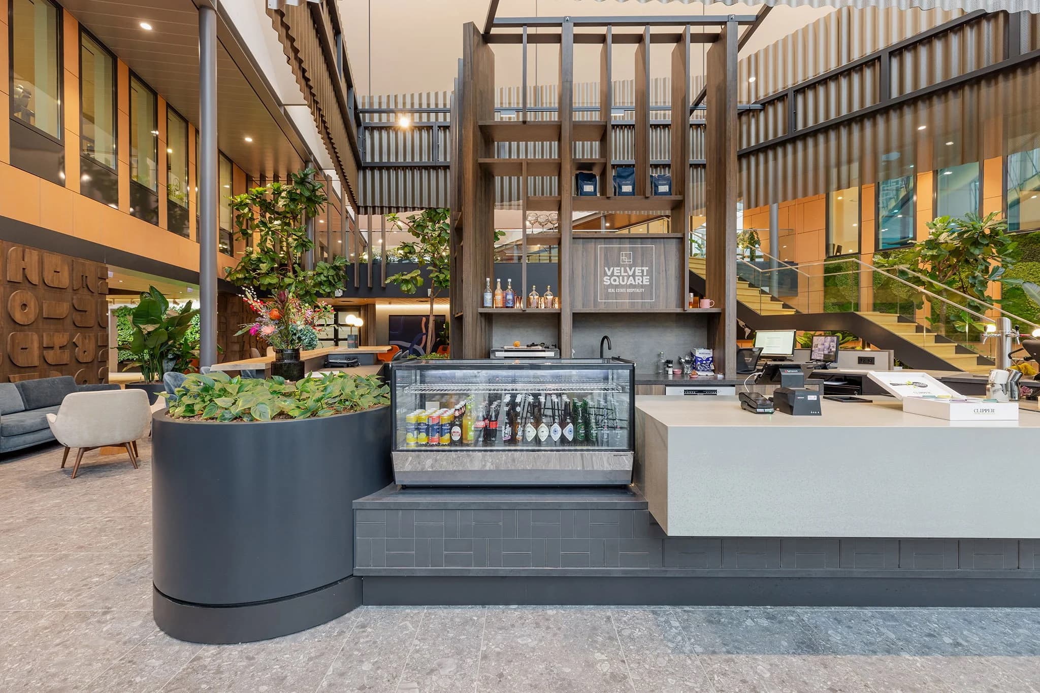 Modern hotel lobby with a sleek reception desk, beverage display, plants, and stylish seating area. Stairs lead to the upper level.