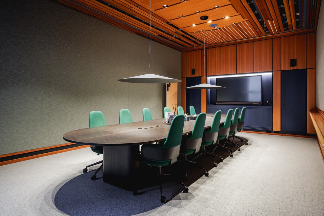 Modern conference room with a long oval table, green chairs, two pendant lights, and a large TV screen on a wood-paneled wall.