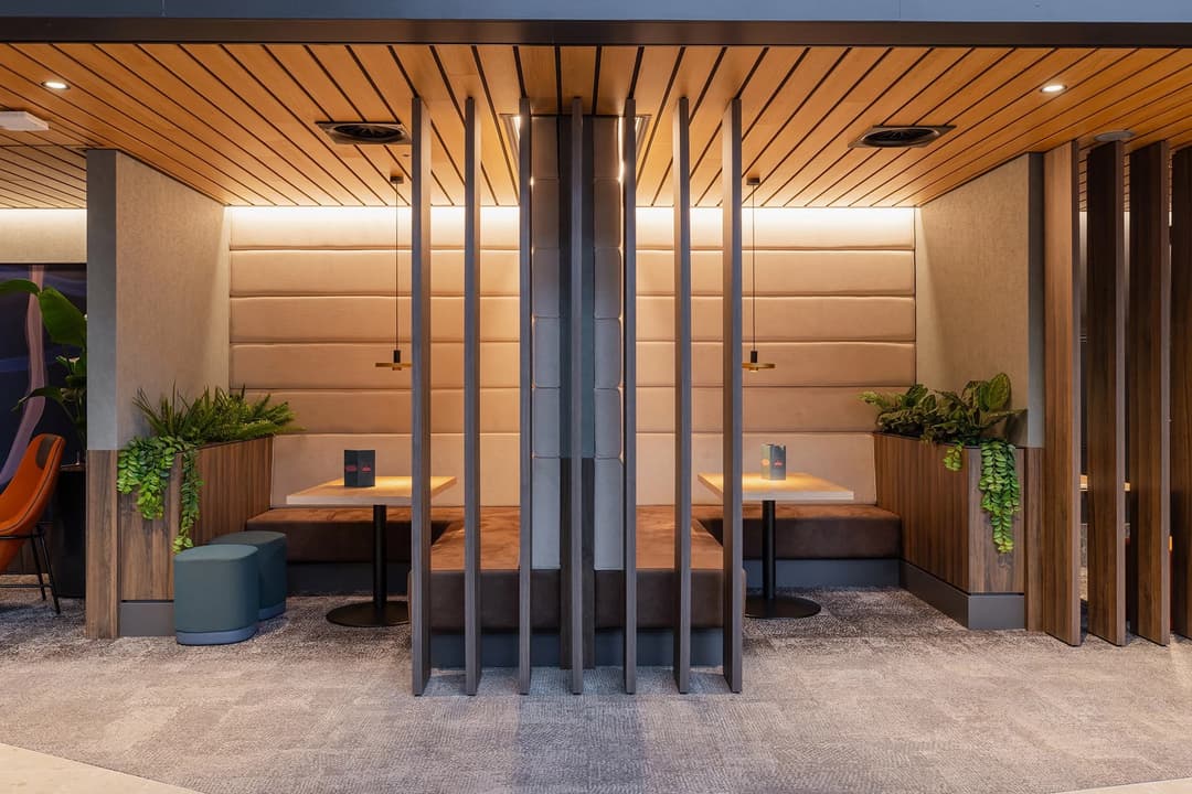 Modern restaurant booths with wooden dividers, beige cushioned seating, plants, and ambient lighting.