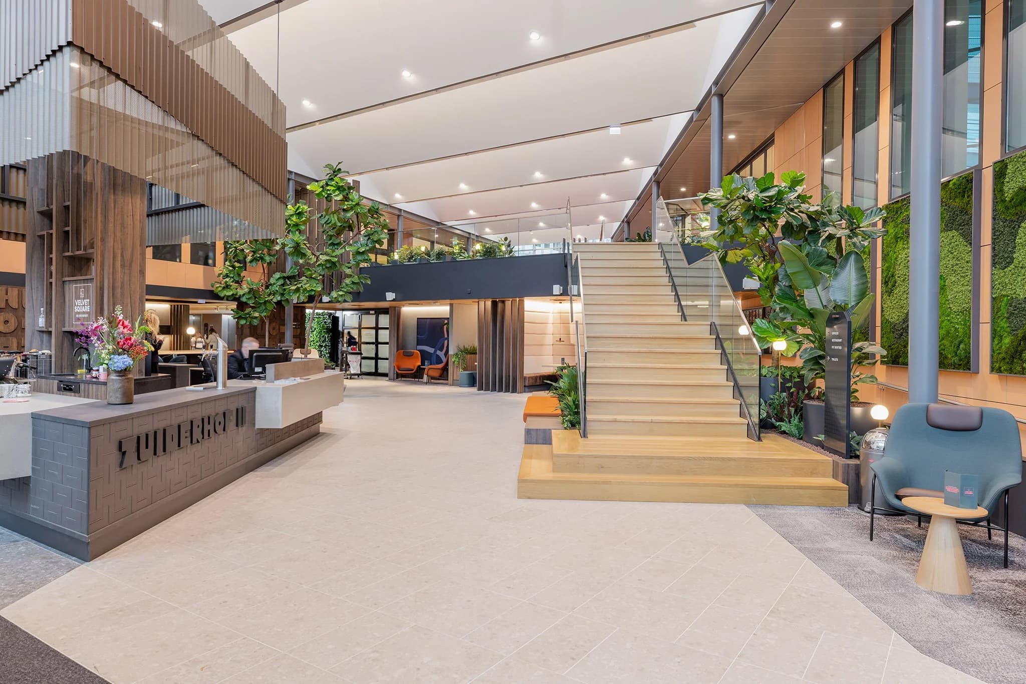 Modern lobby with a large staircase, greenery, seating areas, and a reception desk. Bright lighting and open space create an inviting atmosphere.