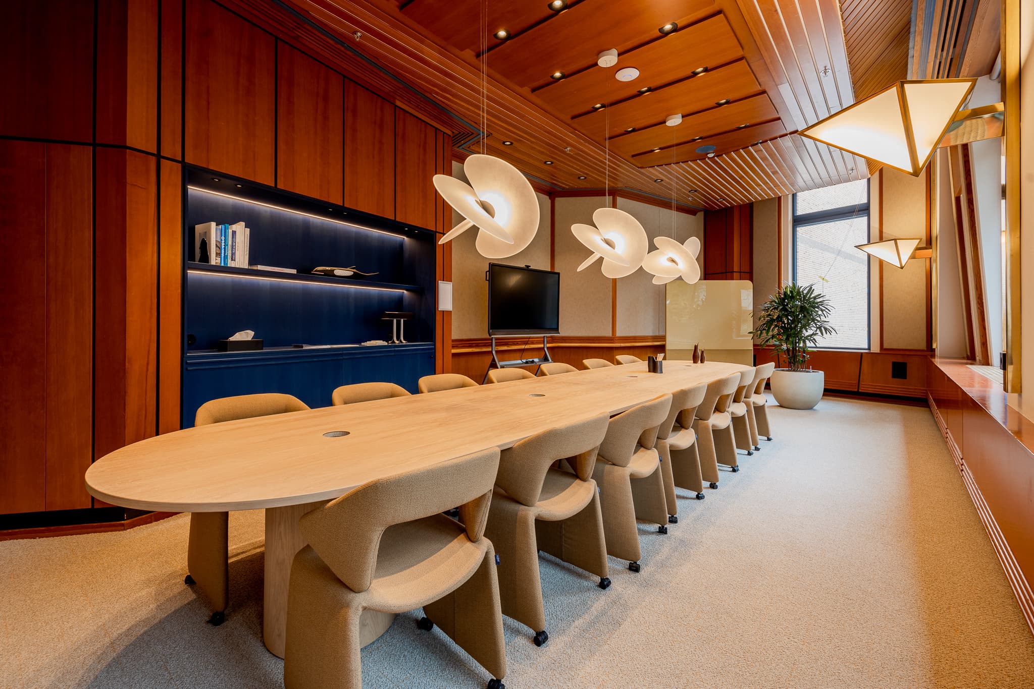 Modern conference room with a long wooden table, beige chairs, unique ceiling lights, wood-paneled walls, and a large plant by the window.
