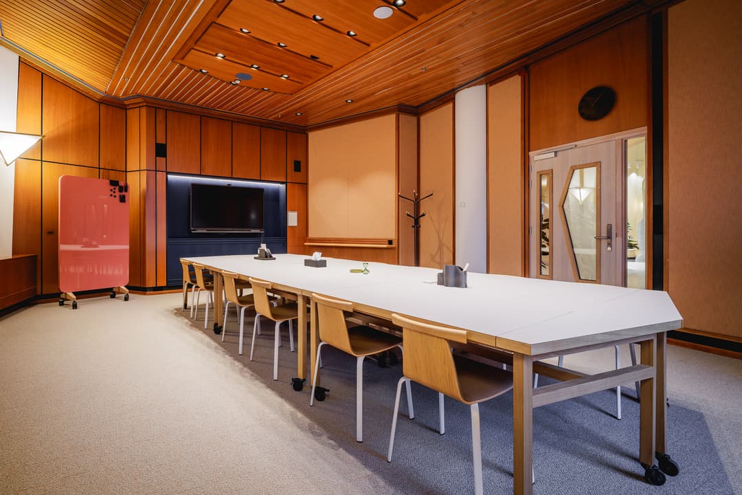 A modern conference room with a long white table, wooden chairs, a wall-mounted TV, and warm wooden paneling. A pink board stands to the left.