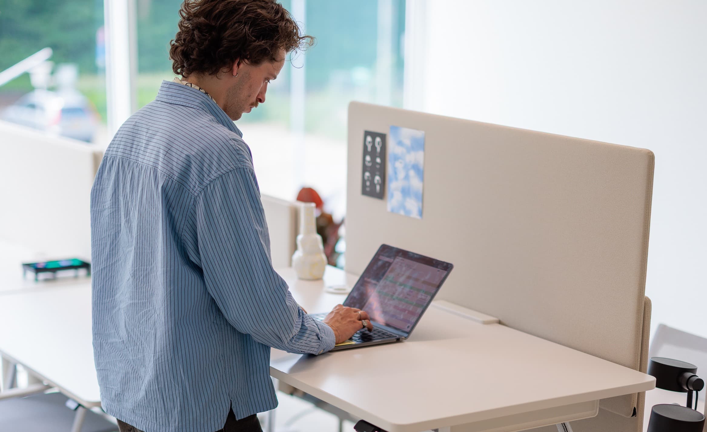 Person in a blue shirt works on a laptop at a standing desk in a bright, modern office space.