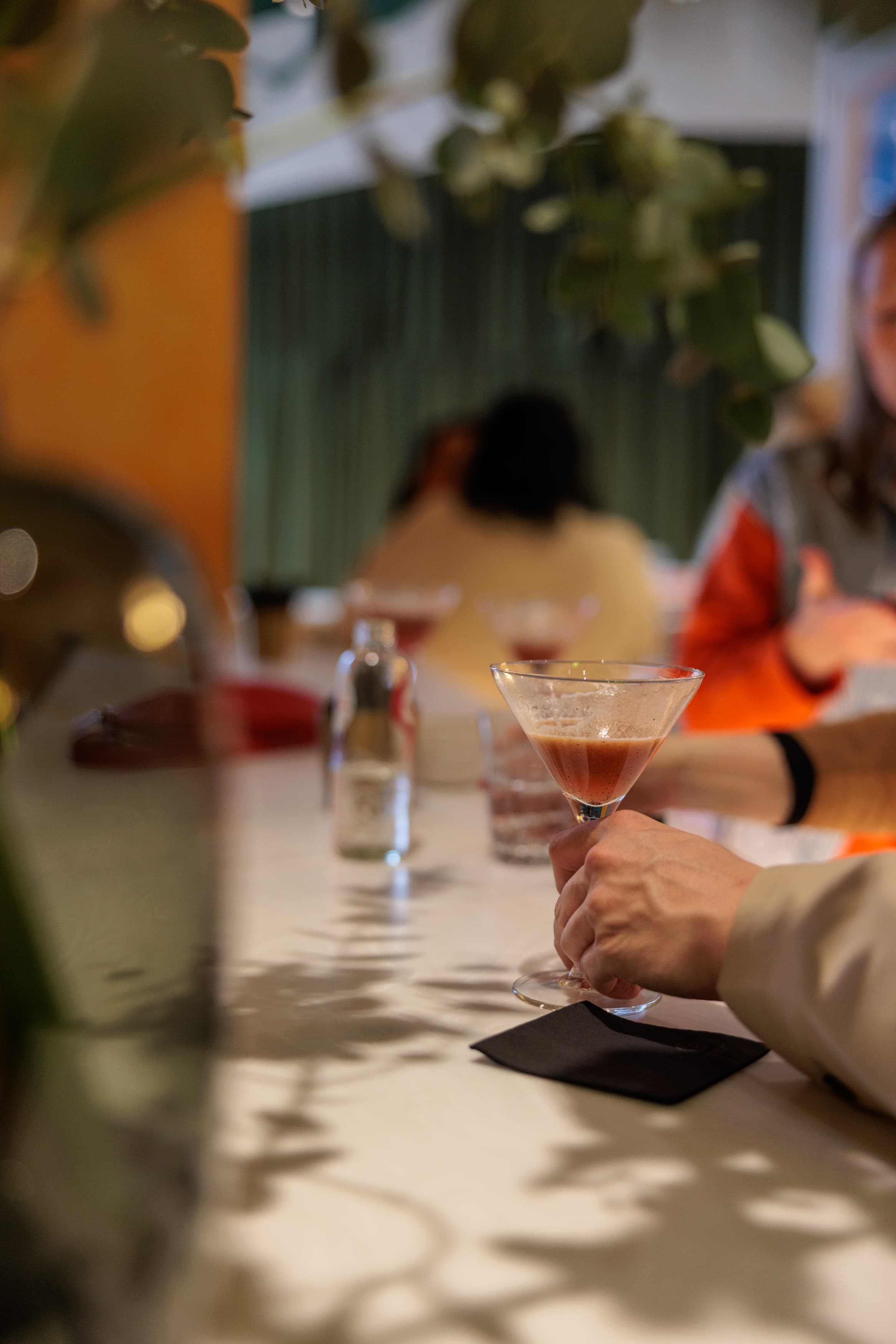 Person holding a cocktail glass at a table in a cozy, dimly lit bar with blurred background and greenery.