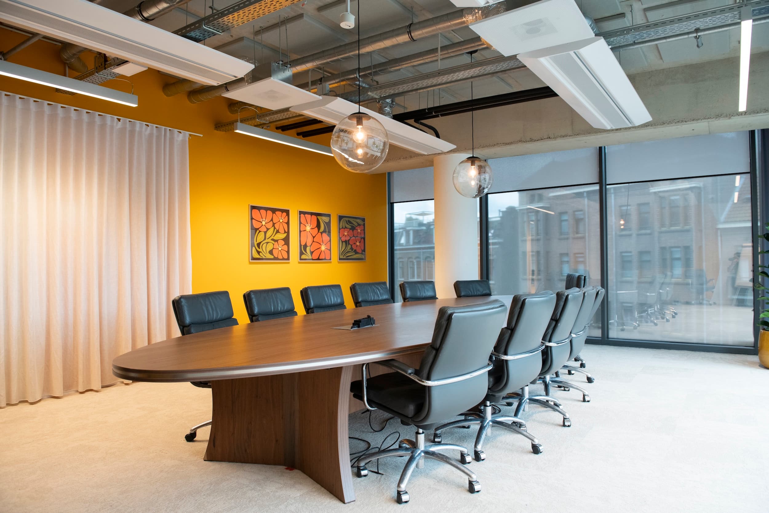 A modern conference room with a long wooden table, black leather chairs, a bright yellow wall, large windows, and ceiling pendant lights.