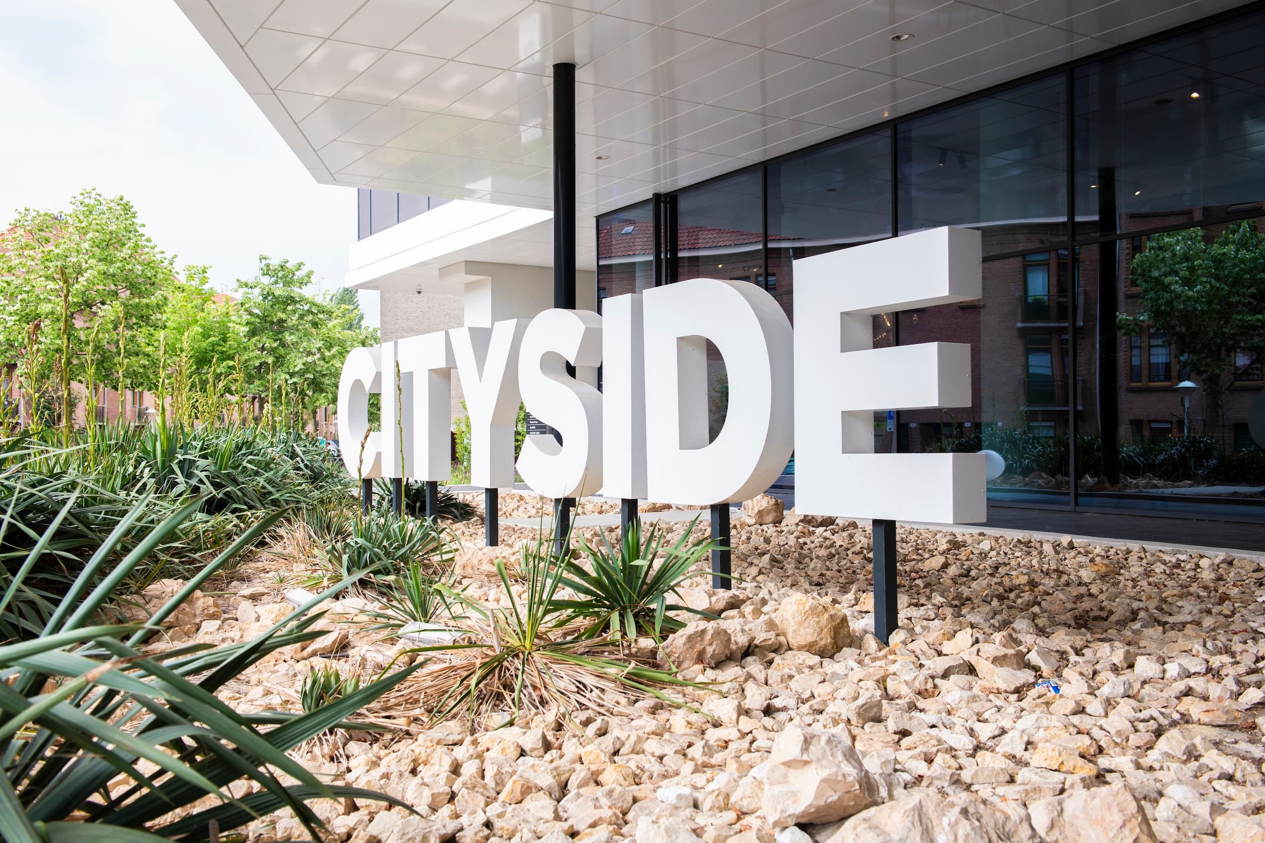 Large white "CITYSIDE" sign on a bed of rocks and plants, with a modern building and trees in the background.