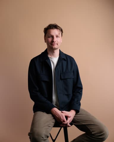 Man sitting on a stool against a beige background, wearing a dark jacket and light pants, hands clasped in front.