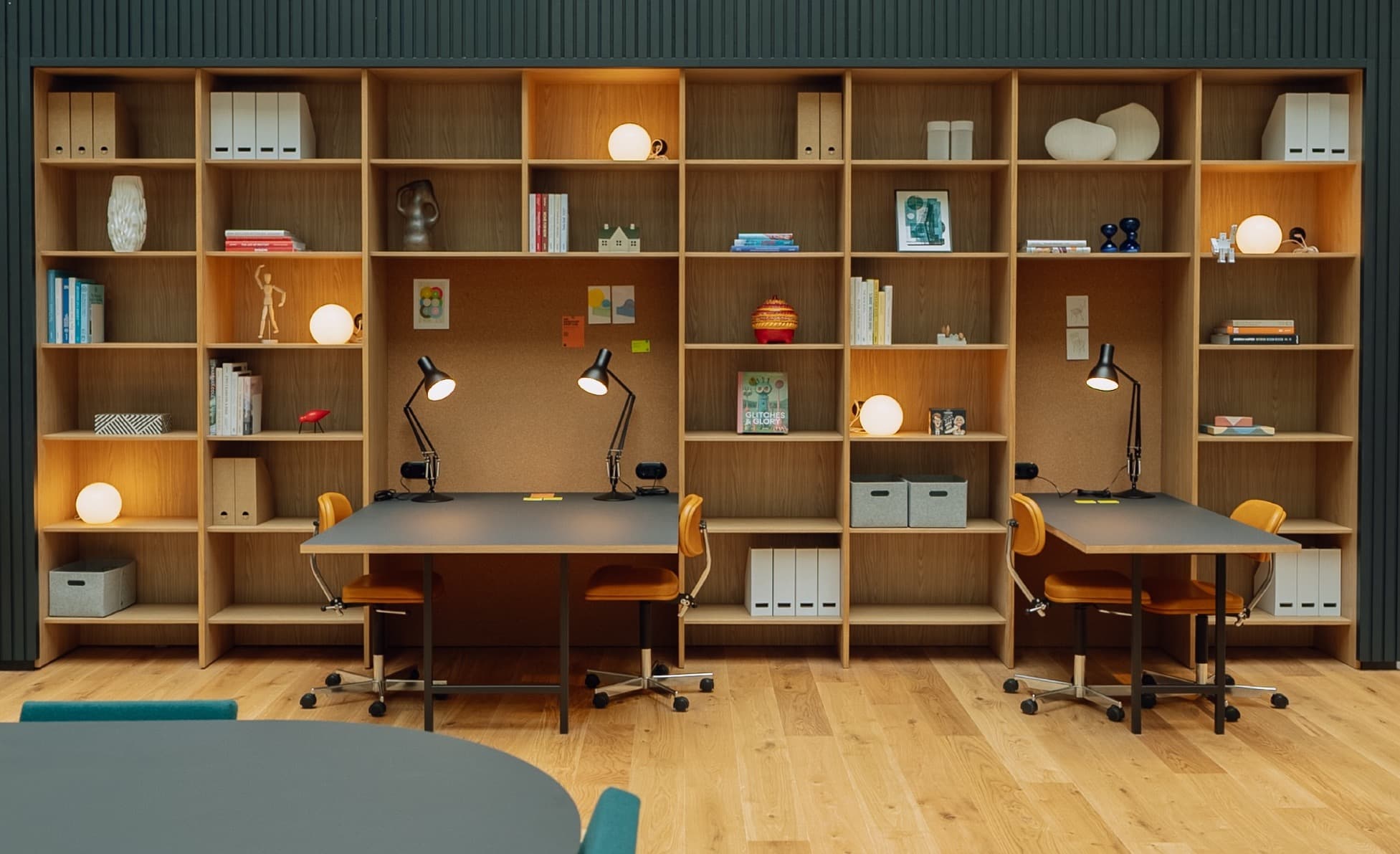 Modern office space with wooden shelves, lamps, and two desks, surrounded by plants. A glass-walled upper floor is visible above.