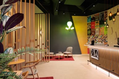 Modern cafe interior with wooden accents, a colorful wall, hanging lights, and a counter with "ZK" signage. Chairs and tables are arranged neatly.