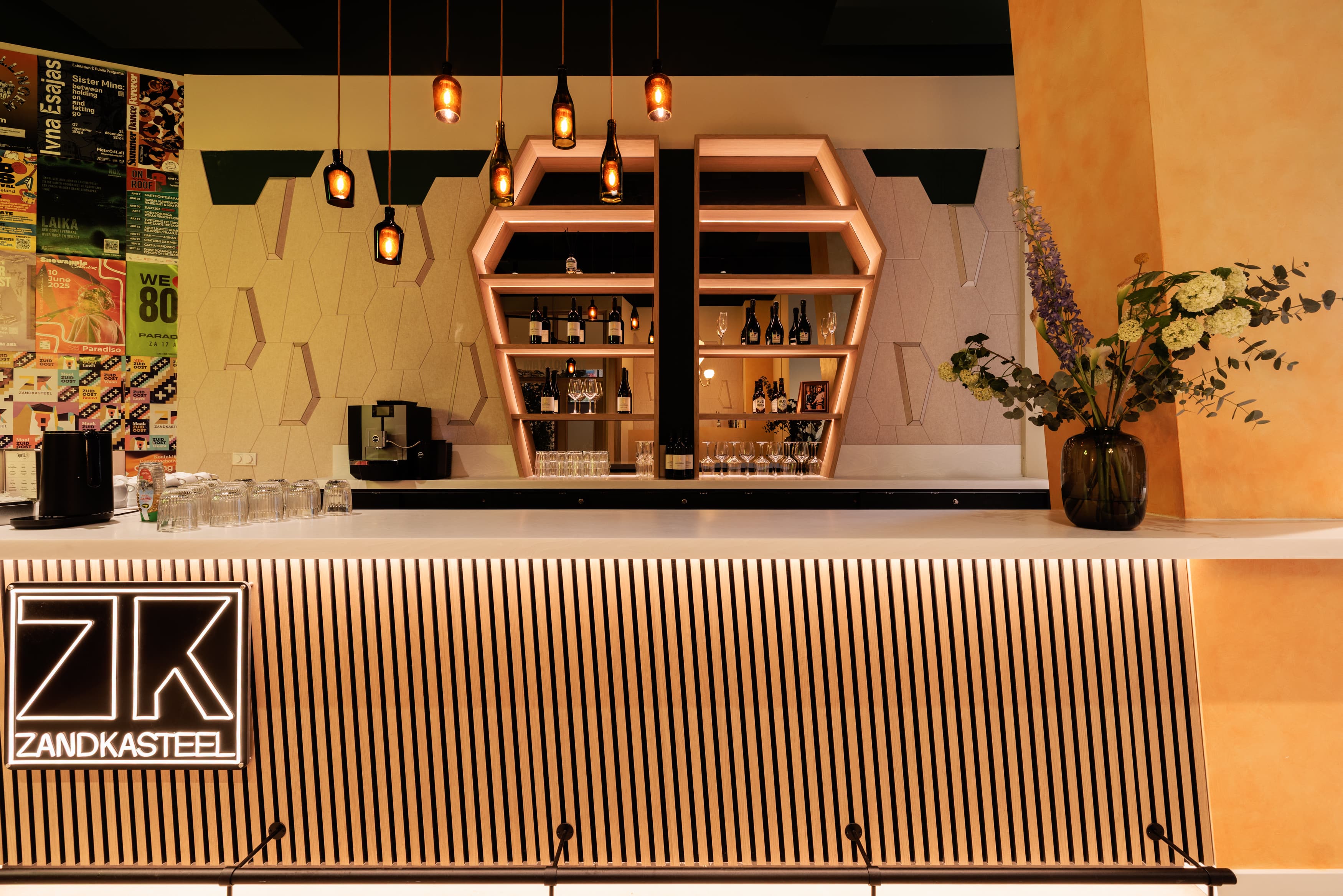 Modern bar interior with geometric shelves, hanging pendant lights, and a vase with flowers. A coffee machine and various bottles are displayed.