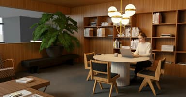 A woman works on a laptop at a round table in a modern, wooden-paneled room with bookshelves, a large plant, and a stylish chandelier.