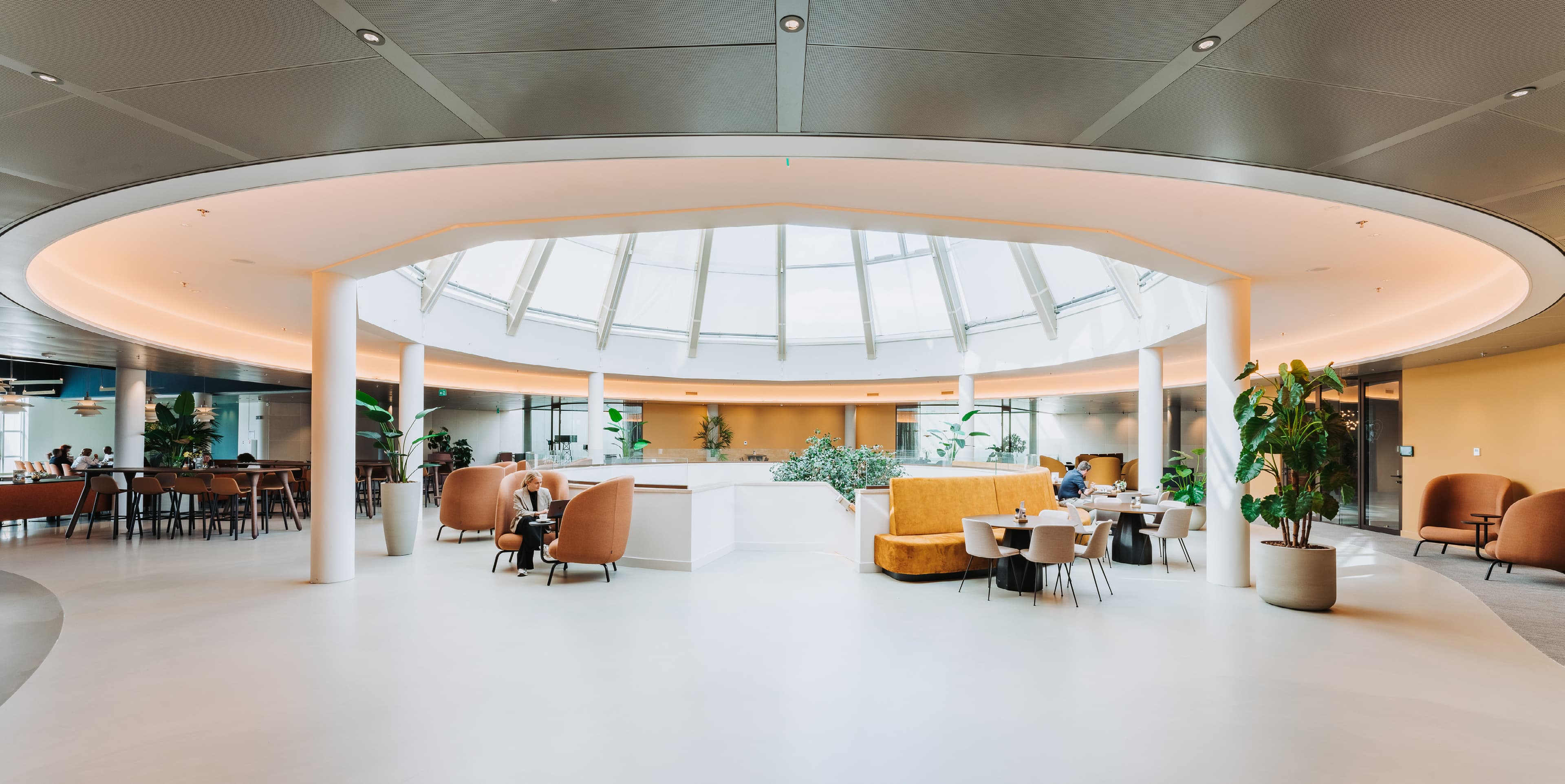 Modern lounge area with circular skylight, orange and beige seating, plants, and tables. Bright and airy atmosphere with a contemporary design.